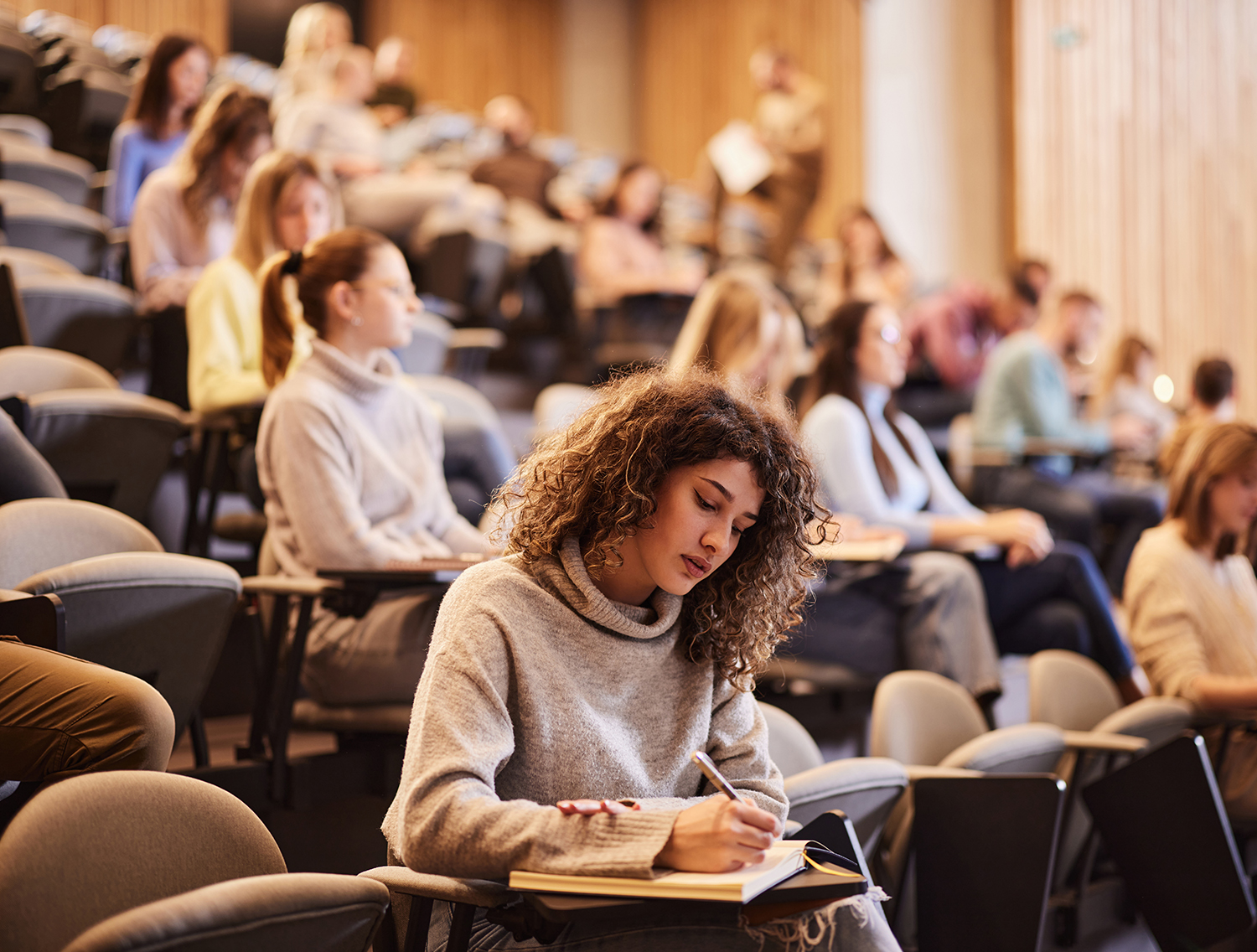 Female college student writing an exam during a class at lecture hall. Her classmate are in the background.