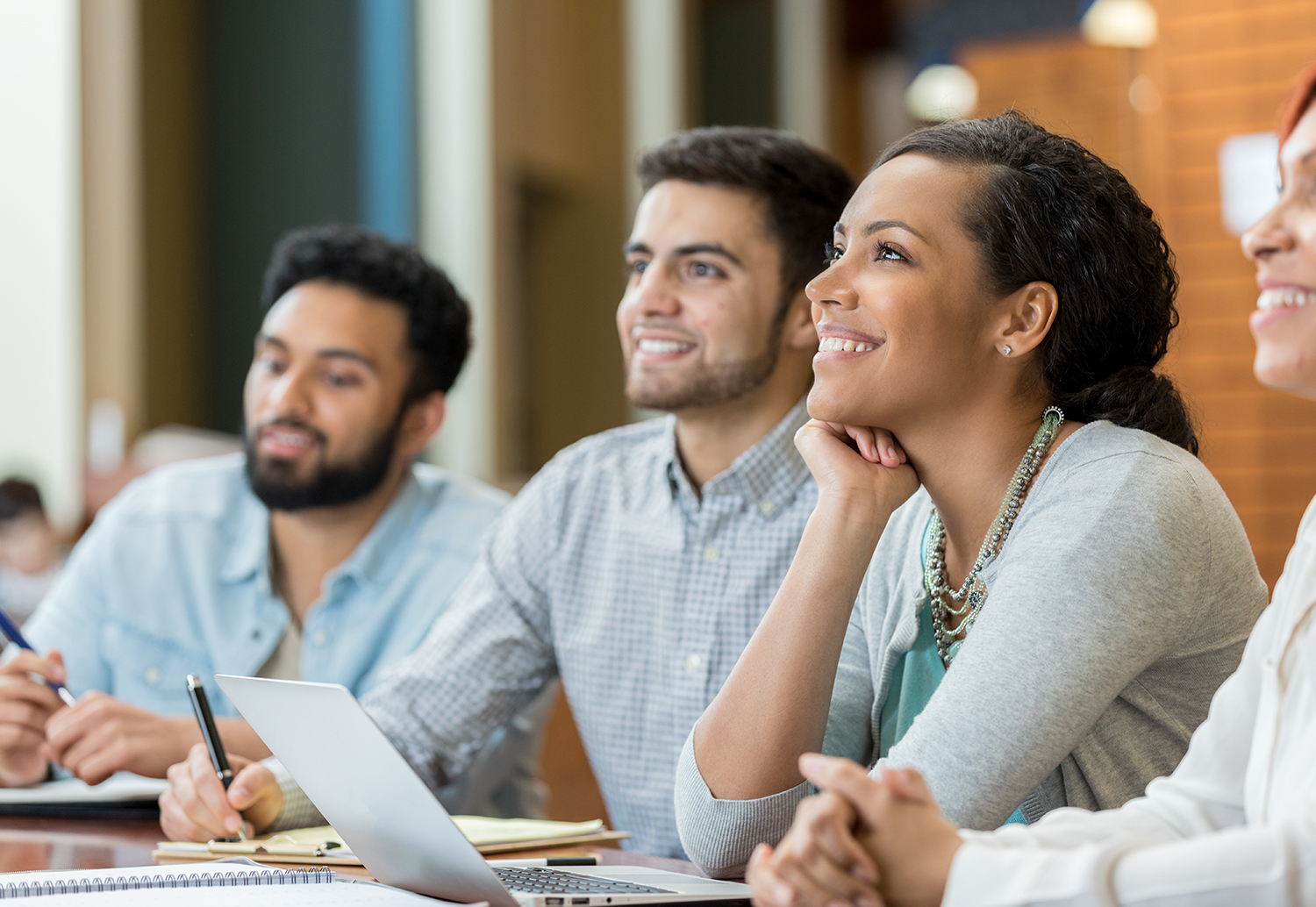 A young woman sits at a table in her university lecture hall with classmates. They all look up with an excited smile as they seem to enjoy the professor's talk.